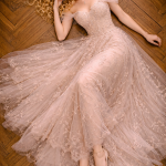 Blonde woman lying on wooden floor in a flowing blush pink lace gown.