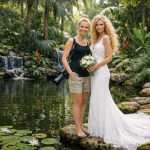 Bride in white lace dress with bouquet standing next to photographer by a pond with lilies and waterfall