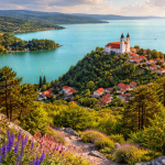 Historic church with red roofs on a lush hill overlooking a turquoise lake with sailboats under a partly cloudy sky.