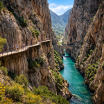 People walking on a narrow wooden walkway along a steep canyon overlooking a turquoise river with a stone bridge.