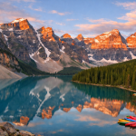 Snowy mountain peaks reflecting in a turquoise lake with colorful kayaks docked on the shore at sunset