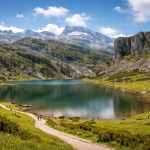 Mountain lake surrounded by grassy hills, rocky cliffs, and distant snow-capped peaks under a partly cloudy sky.