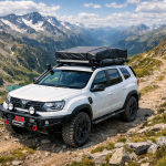 White 4x4 SUV with rooftop tent on rocky mountain trail with snow-covered peaks and valley behind.