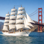 White tall sailing ship with full sails near the red Golden Gate Bridge on a sunny day.