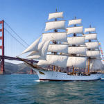 Large white tall ship with full sails sailing near the Golden Gate Bridge on clear water.