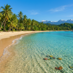 Clear turquoise water, sandy beach with palm trees, and snow-capped mountains in the distance.