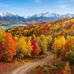 Winding dirt road through colorful autumn trees with snow-capped mountains and a blue lake in the background.